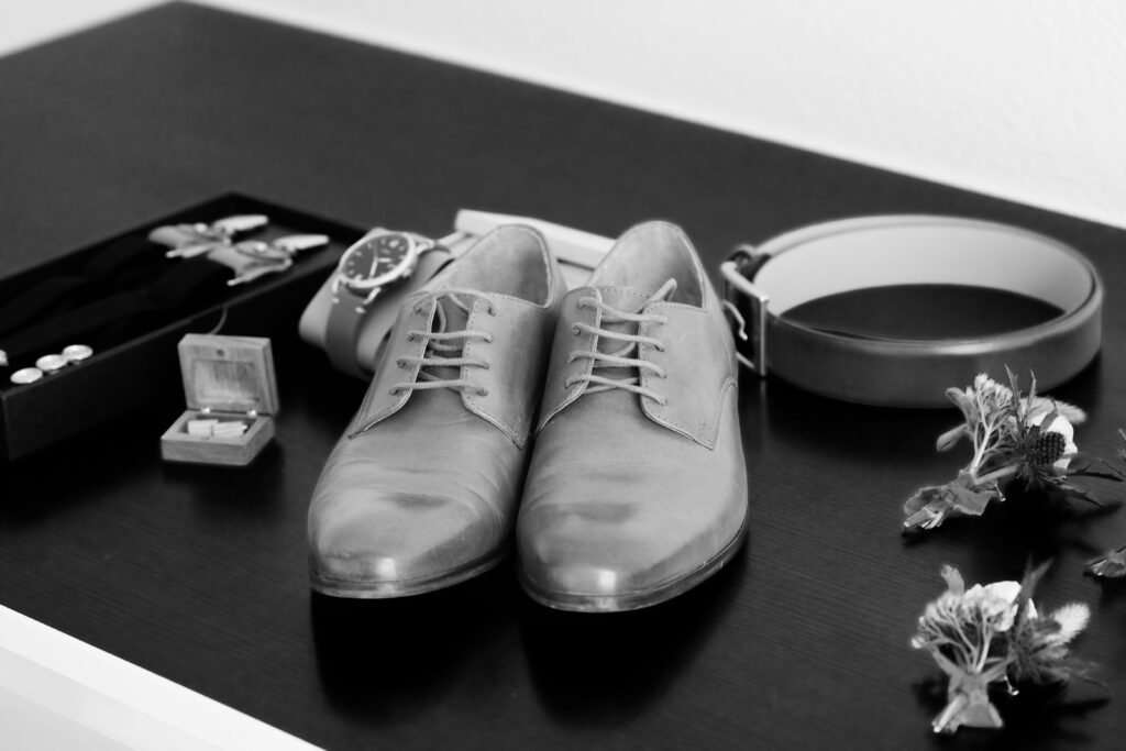 Détails des préparatifs du marié en noir et blanc avec chaussures en cuir, montre, ceinture, bretelles et boutonnières fleuries.