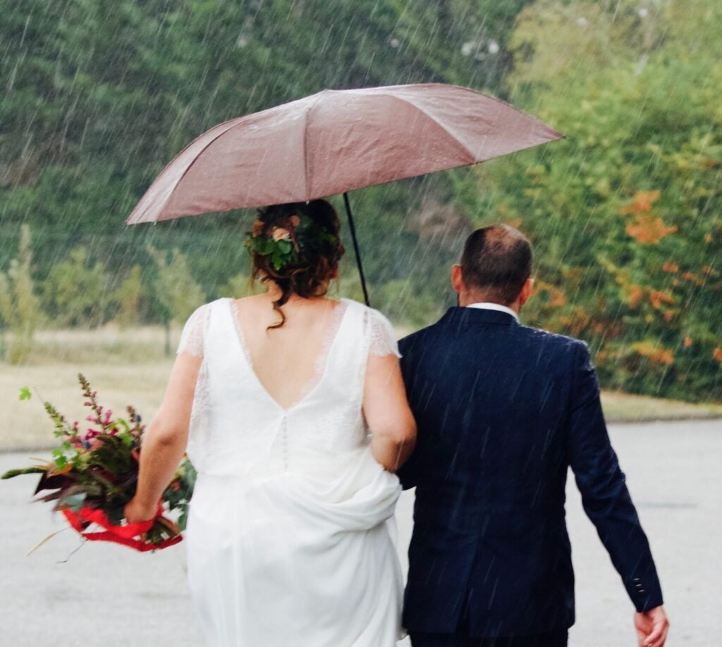 Mariés marchant main dans la main sous la pluie, à l'abri sous un parapluie. Cette image illustre l'importance d'un plan B bien orchestré lors de l'organisation d'un mariage en extérieur par une wedding planner en Seine-et-Marne, transformant un imprévu météorologique en un moment intime et romantique. La robe de la mariée vole légèrement au vent.